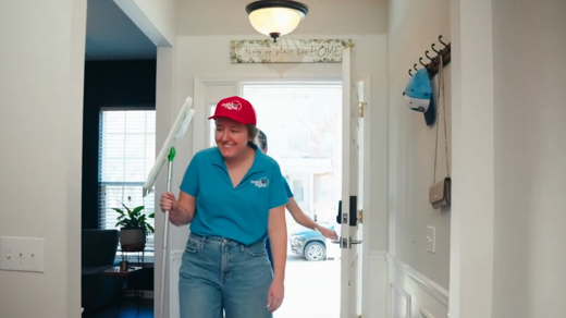 a woman in a ball cap walking into a home with cleaning supplies